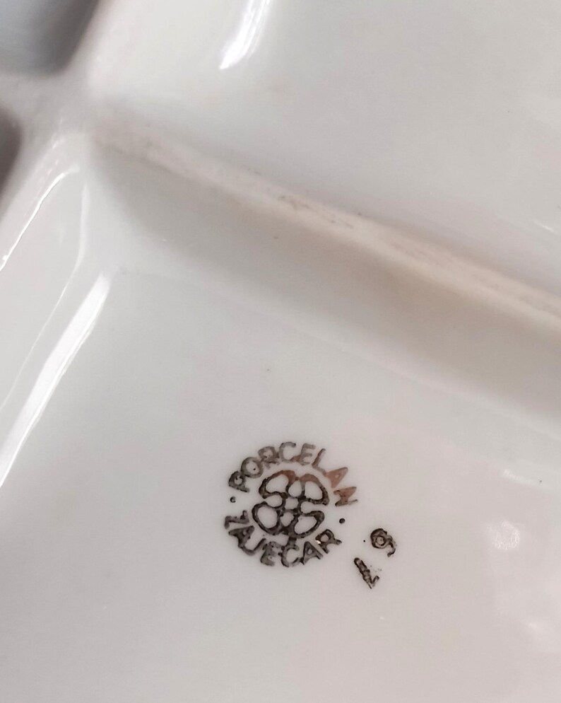 May include: White porcelain serving dish with multiple compartments. The dish has a glossy finish and a decorative emblem with the text "PORCELAN ZUECAR" in black. The number "6" is also visible.