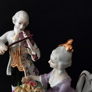May include: A porcelain figurine depicting a man playing a violin and a woman seated nearby. The man wears a white coat with gold accents, while the woman is in a purple dress. The figurine has a black background.
