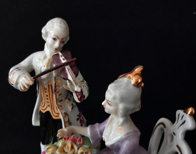 May include: A porcelain figurine depicting a man playing a violin and a woman seated nearby. The man wears a white coat with gold accents, while the woman is in a purple dress. The figurine has a black background.