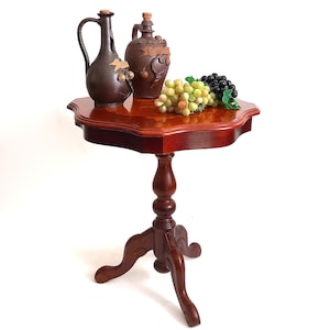Peut inclure: Petite table d'appoint en bois foncé avec un plateau festonné, supportant deux cruches en céramique marron et une grappe de raisin. La table a un pied en colonne et trois pieds incurvés.