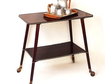 May include: A dark brown wooden serving cart with a glass top and a lower shelf. The cart has four legs and gold-colored wheels. A silver tea set is on the top shelf.