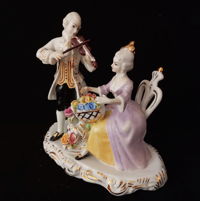 May include: A porcelain figurine depicting a man playing a violin and a woman seated with a basket of flowers. The man wears a white coat with gold trim, black pants, and the woman wears a lavender dress. The base is white with gold accents.