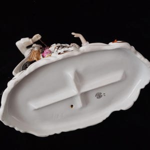 May include: A porcelain figurine depicting a man and woman in period clothing. The base is white with a scalloped edge. The man is wearing a white coat with gold accents, and the woman is wearing a white dress. The figurine has a black background.