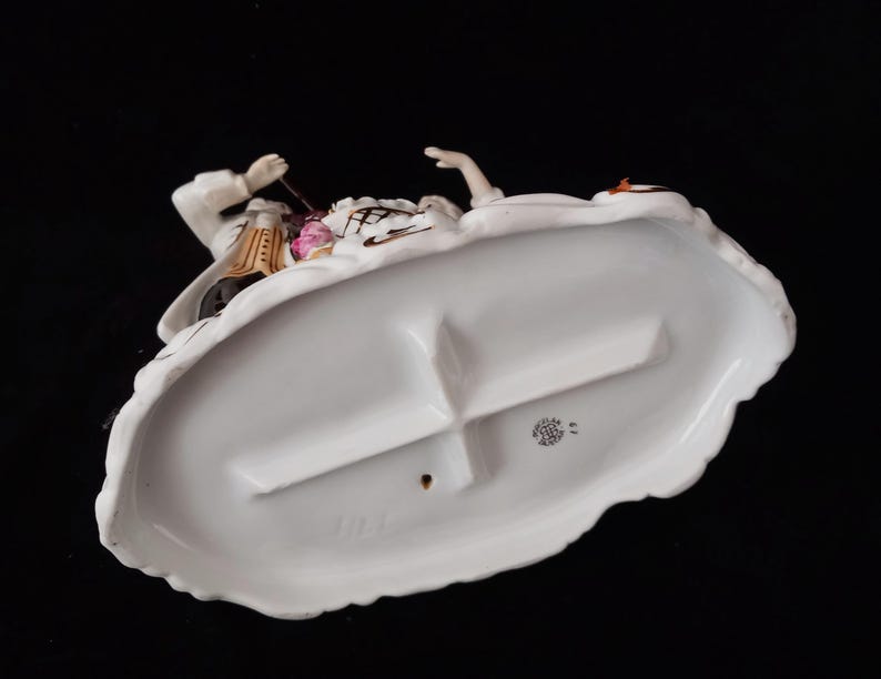 May include: A porcelain figurine depicting a man and woman in period clothing. The base is white with a scalloped edge. The man is wearing a white coat with gold accents, and the woman is wearing a white dress. The figurine has a black background.