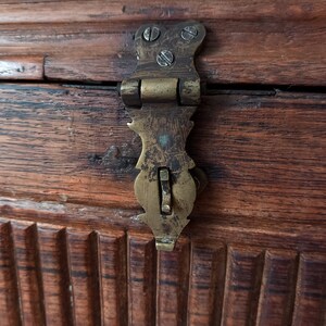 May include: A close-up of a brass latch on a wooden chest. The latch is ornate and has a small, rectangular lever.
