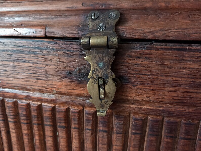 May include: A close-up of a brass latch on a wooden chest. The latch is ornate and has a small, rectangular lever.