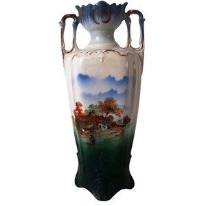 May include: A white porcelain vase with blue and gold accents. The vase features a painted landscape scene with a cottage, trees, and clouds. The vase has two handles and a wide mouth.