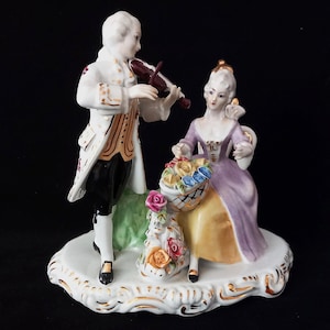 May include: A porcelain figurine depicting a man playing a violin and a woman seated with a basket of flowers. The man wears a white coat with gold trim, black pants, and a wig. The woman wears a purple dress. The base is white with gold accents.