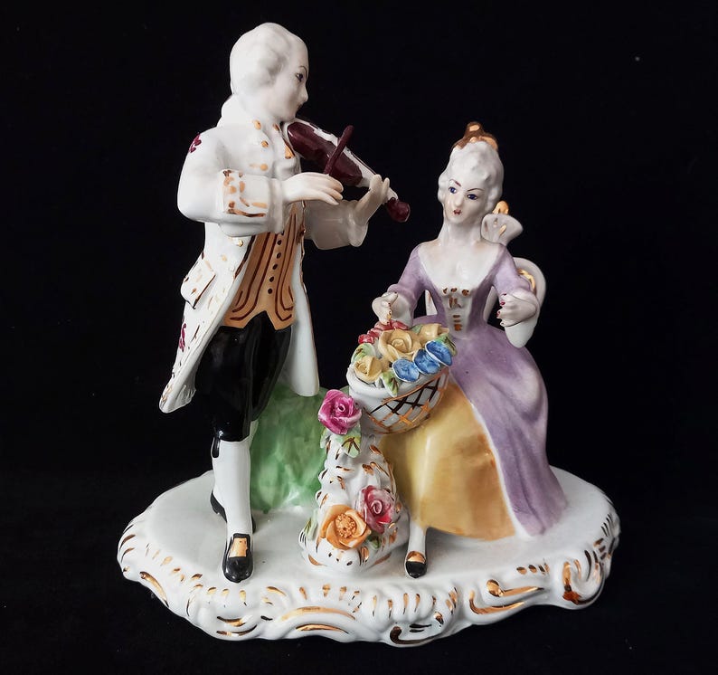 May include: A porcelain figurine depicting a man playing a violin and a woman seated with a basket of flowers. The man wears a white coat with gold trim, black pants, and a wig. The woman wears a purple dress. The base is white with gold accents.