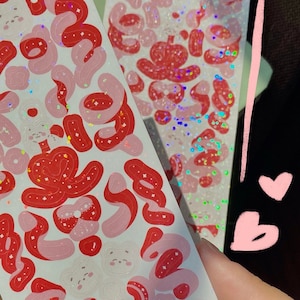 May include: Two sheets of pink and red holographic stickers with a variety of shapes and designs. The stickers are on a white background.