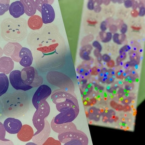 May include: A sheet of stickers featuring a variety of cute designs, including bears, flowers, and swirls. The stickers are in shades of purple, pink, and white. The sheet is on a green surface.