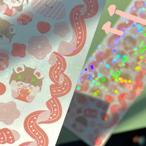 May include: A sheet of pink and white stickers with a variety of cute cartoon characters, hearts, and bows. The stickers are arranged in a diagonal pattern on a white background. The sheet is partially obscured by a sheet of iridescent holographic stickers.