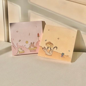 May include: Two pastel colored sticky note pads with cute illustrations. The pink pad features a rabbit and a mouse playing in a wave. The yellow pad features a cat carrying a pastry and a mouse watching.