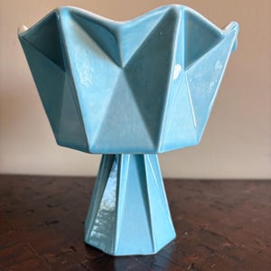 Könnte beinhalten: Eine hellblaue Keramik-Sockelschale mit geometrischem, facettiertem Design. Die Schale hat einen breiten, gewellten Rand und einen sechseckigen Sockel. Die Schale ist etwa 20 cm hoch.