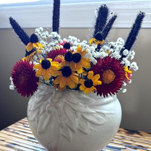 Pode incluir: Um vaso de cerâmica branco com um desenho floral contém um arranjo floral vibrante. O buquê inclui margaridas amarelas e vermelhas, gipsofila branca e caules altos pretos semelhantes a grama. O vaso está sobre uma mesa de bambu.