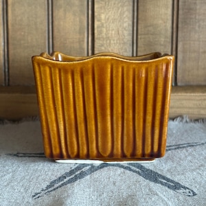 Può includere: Piccolo vaso rettangolare in ceramica color ambra caldo, con un design a coste. Il vaso ha un bordo superiore smerlato e una base bianca. Poggia su un tessuto fantasia con bordo sfrangiato, su uno sfondo in legno.
