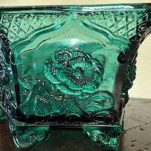 Puede incluir: Una jardinera cuadrada de vidrio verde esmeralda con detalles florales y geométricos ornamentados. La jardinera presenta un diseño floral en relieve en la parte delantera y los lados, con un patrón de diamantes en los bordes superiores. Descansa sobre cuatro patas pequeñas.
