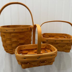 May include: Three woven wooden baskets in varying sizes. The baskets are a warm, golden-brown colour and feature arched handles. The largest basket is round, while the other two are rectangular. Each basket is constructed with a woven pattern.