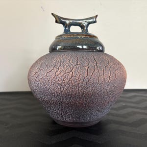 Pode incluir: Um pote de cerâmica com um esmalte texturizado marrom e roxo. A tampa tem um design único e abstrato com um esmalte azul escuro.