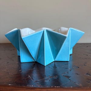 May include: A geometric ceramic bowl with a folded paper-like design. The exterior is a glossy, light blue, while the interior is white. The bowl sits on a dark wooden surface, showcasing its angular form and contrasting colors.