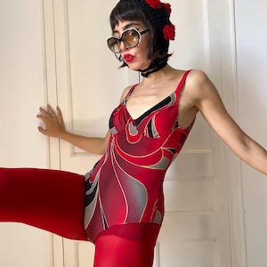 Op de afbeelding: Een badpak uit één stuk met een abstract wervelpatroon in rood, zwart en grijs met dunne schouderbandjes. Het kledingstuk wordt gecombineerd met een effen rode panty, een zwart gehaakt hoofddeksel met rode bloemen en een ronde zonnebril.