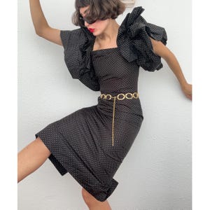 May include: A black, knee-length dress with white polka dots, a square neckline, and large, ruffled sleeves. A gold chain belt cinches the waist. The dress is a vintage style.