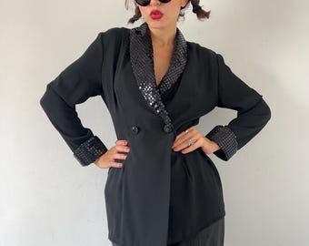 Puede incluir: Blazer negro con solapas y puños con lentejuelas. La chaqueta cruzada tiene dos botones negros y mangas largas. El blazer se usa con pantalones oscuros. Las solapas y los puños tienen un patrón a cuadros.