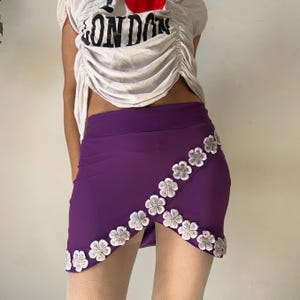 Pode incluir: Uma minissaia roxa com uma borda frontal diagonal estilo envelope, adornada com uma fileira de aplicações de renda floral branca. A saia tem uma cor base roxa sólida e uma bainha que segue a linha diagonal dos detalhes florais brancos.