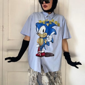 Op de afbeelding: Een lichtblauw overhemd met korte mouwen en knoopsluiting, voorzien van een grote grafische print van het personage Sonic the Hedgehog op de voorzijde. De afbeelding bevat gele Koreaanse tekst boven het personage. Het overhemd heeft een kraag.