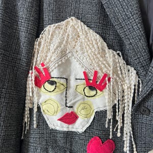 May include: A dark grey plaid blazer featuring a large fabric applique of a face. The face has cream lace hair, bright red felt eyelashes and lips, yellow painted cheeks and eyes, and black embroidered outlines. A red felt heart sits below.