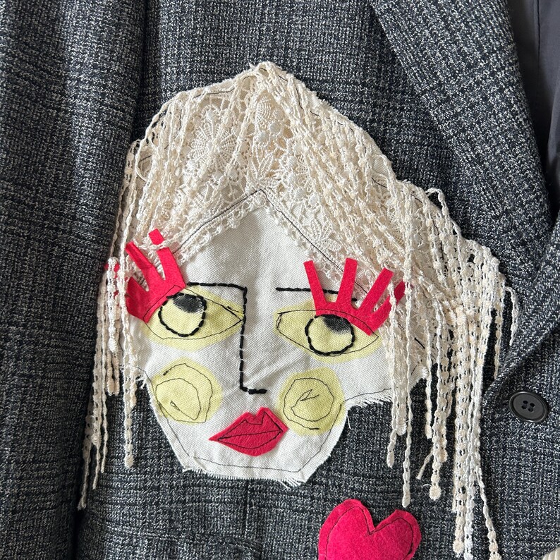 May include: A dark grey plaid blazer featuring a large fabric applique of a face. The face has cream lace hair, bright red felt eyelashes and lips, yellow painted cheeks and eyes, and black embroidered outlines. A red felt heart sits below.