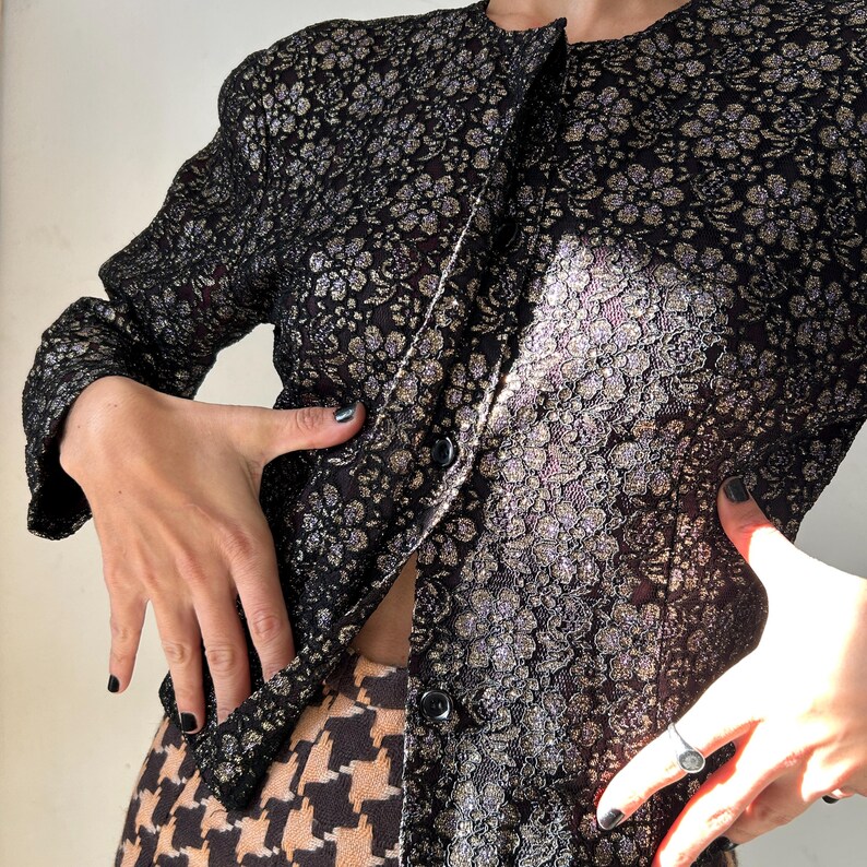 May include: A long-sleeved jacket featuring a black and metallic gold floral pattern. The garment has a round neckline, a front button closure with two visible black buttons, and a textured fabric construction with a subtle shimmer throughout.