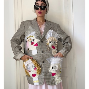 Könnte beinhalten: Ein braun-weiß karierter Blazer mit vier verschiedenen Stoffapplikationen in Form von Gesichtern, verziert mit Stickereien, Spitzenakzenten und goldenen Pailletten. Der Blazer hat ein steigendes Revers, eine Knopfleiste mit drei Knöpfen und Ärmel mit Umschlag.