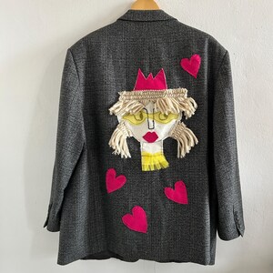 May include: A dark gray plaid blazer featuring a large, central fabric appliqu&eacute; of a face with yellow glasses, red lips, and a pink crown. The design includes cream-colored yarn hair and four bright pink fabric hearts scattered around the face.