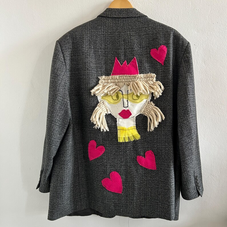 May include: A dark gray plaid blazer featuring a large, central fabric appliqu&eacute; of a face with yellow glasses, red lips, and a pink crown. The design includes cream-colored yarn hair and four bright pink fabric hearts scattered around the face.