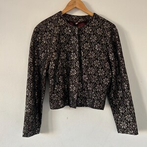 May include: A long-sleeved jacket featuring a dark brown base with a metallic silver floral pattern throughout. The garment has a round neckline, a straight hem, and a front closure, displayed on a wooden hanger against a plain background.