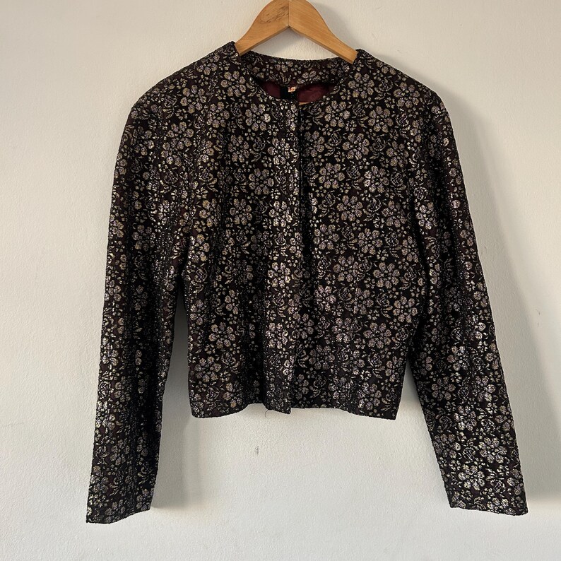 May include: A long-sleeved jacket featuring a dark brown base with a metallic silver floral pattern throughout. The garment has a round neckline, a straight hem, and a front closure, displayed on a wooden hanger against a plain background.