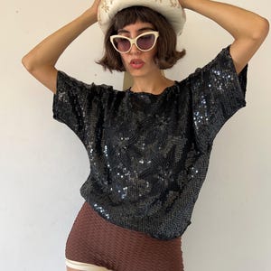 May include: A black short-sleeved top covered in dense, shimmering sequins arranged in a floral pattern. The garment features a round neckline and a loose, boxy fit. The sequins reflect light, creating a textured, high-shine surface across the fabric.