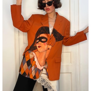 Könnte beinhalten: Ein orangefarbener Blazer mit einer handgemalten Illustration einer Figur, die eine schwarze Maske und ein Kleidungsstück mit Harlekinmuster in Orange-, Blau- und Brauntönen trägt. Der Blazer hat lange Ärmel, ein Revers und zwei Pattentaschen.