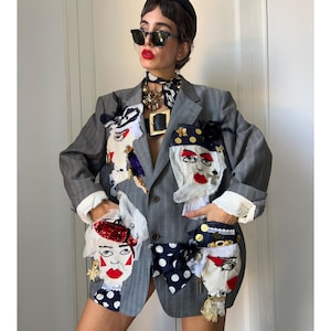 Op de afbeelding: Een grijze blazer met krijtstreep voorzien van meerdere stoffen gezichtapplicaties met rode lippen, zwart-witte stippen en goudkleurige versieringen. De blazer heeft een reverskraag en lange mouwen met witte manchetten en een knoopsluiting.