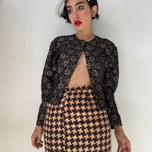 May include: A long-sleeved jacket featuring a black and silver floral pattern paired with a high-waisted skirt displaying a tan and black houndstooth print. The jacket has a round neckline and is worn open, while the skirt has a straight silhouette.