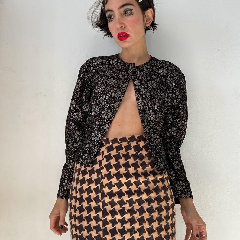 May include: A long-sleeved jacket featuring a black and silver floral pattern paired with a high-waisted skirt displaying a tan and black houndstooth print. The jacket has a round neckline and is worn open, while the skirt has a straight silhouette.