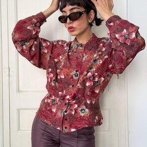 May include: A button-down blouse with a floral pattern in shades of red, pink, and white. The blouse has long, puffy sleeves and a fitted waist. The floral design is set against a paisley and dotted background. The model is wearing sunglasses.