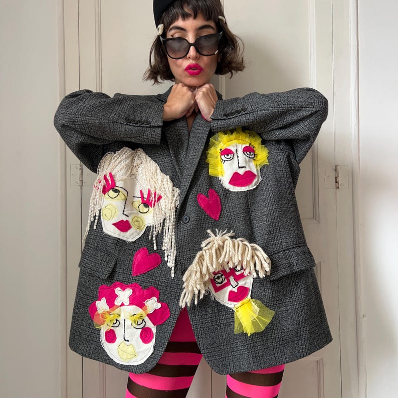 May include: A charcoal gray plaid blazer featuring multiple hand-stitched fabric face appliqu&eacute;s with yarn hair, tulle accents, and bright pink heart patches. The blazer has a structured collar, long sleeves, and two front flap pockets.