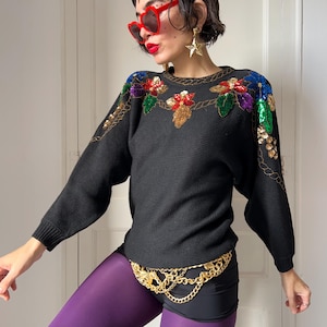 May include: Black sweater with floral sequin detailing in red, gold, green, and blue. The sweater has long sleeves and a gold chain belt. The sweater is a vintage style.