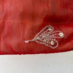 May include: A vibrant red, semi-sheer fabric featuring an intricate silver-toned metallic embellishment. The decorative element consists of multiple interconnected chain-like loops arranged in a fan shape, accented with small, circular silver discs.