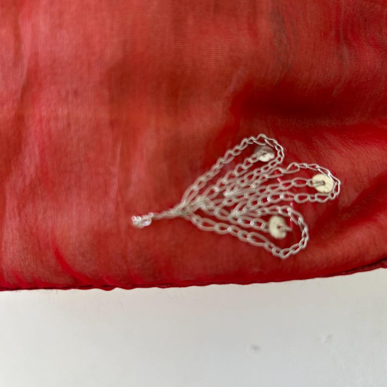 May include: A vibrant red, semi-sheer fabric featuring an intricate silver-toned metallic embellishment. The decorative element consists of multiple interconnected chain-like loops arranged in a fan shape, accented with small, circular silver discs.