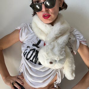 May include: A fluffy white scarf shaped like a bunny, with a plush head and long ears. The scarf is made of soft, faux fur material. The person in the image is wearing a white t-shirt with black text and sunglasses.