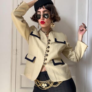 Pode incluir: Um blazer bege com acabamento preto nos bolsos e na gola, apresentando uma fileira de botões redondos estampados na frente. O blazer é combinado com calças pretas, um cinto de corrente dourada com um pingente de folha, brincos de argola dourados e óculos de sol.