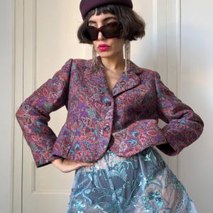 May include: A cropped blazer featuring a multi-colored paisley pattern in shades of red, purple, and teal with two dark buttons. Paired with sheer, light-colored trousers adorned with intricate teal sequin floral embroidery and embellishments.